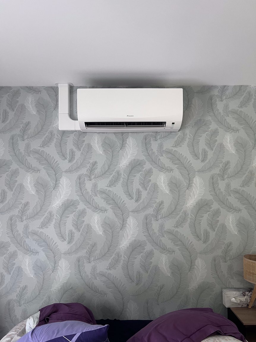 Unité intérieure Daikin posée au-dessus du lit dans une chambre, goulotte blanche descendant à la verticale